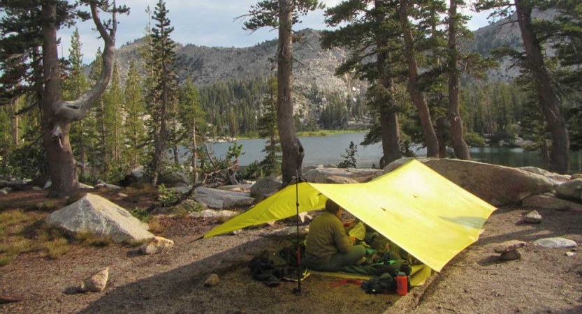 A yellow tarp shelter rests beside a body of water surrounded by trees. Behind the water there are mountains. 